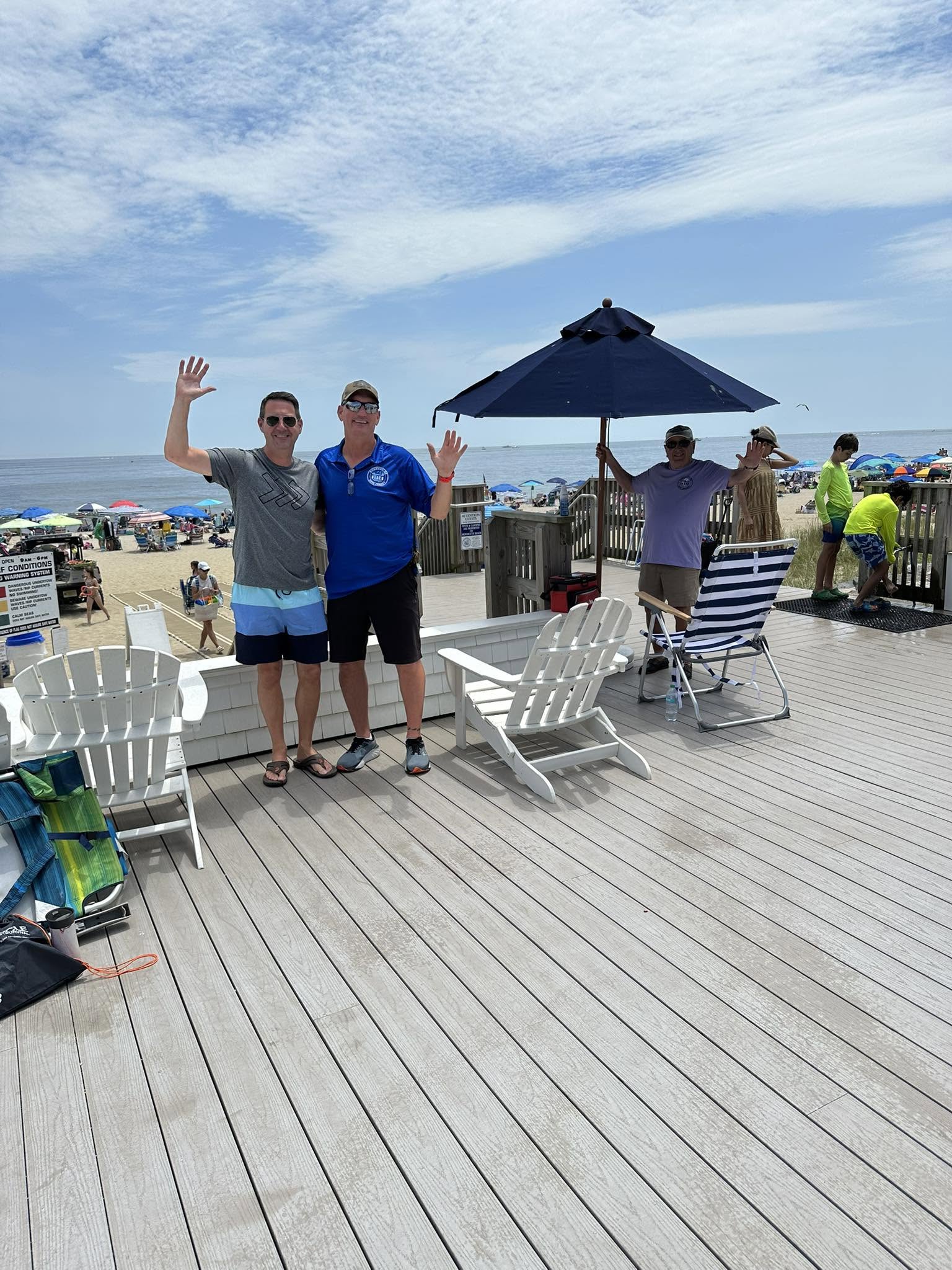 Beachfront deck overlooking the Atlantic Ocean in Monmouth Beach, New Jersey, with white Adirondack chairs, a large umbrella, visitors enjoying the waterfront, and a busy sandy beach filled with colorful umbrellas.
