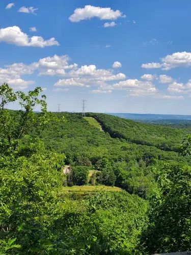 Scenic overlook in Montville, New Jersey, with an expansive view across densely forested green hills, rolling terrain, and a distant horizon beneath a bright blue sky dotted with white clouds.