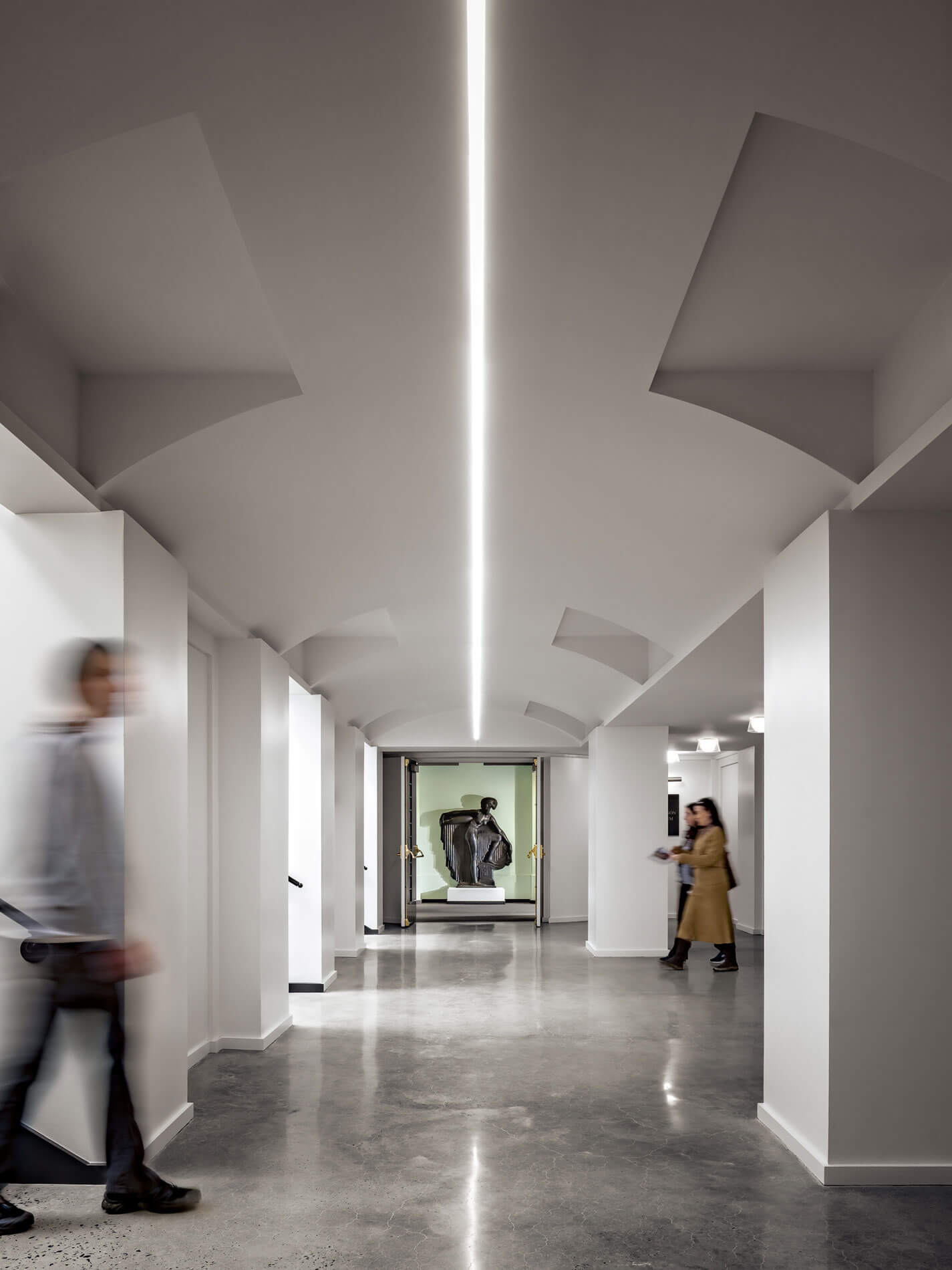 Interior corridor of the Newark Museum of Art in Newark, New Jersey, featuring clean contemporary architecture, vaulted white ceilings, polished concrete floors, and visitors moving through the gallery space. A sculpture displayed at the end of the hallway highlights the museum's collection of art and cultural exhibits.