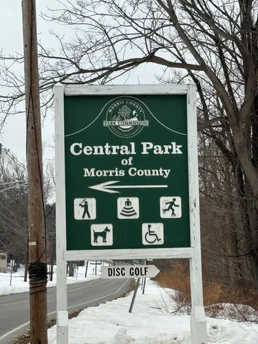 A roadside sign for Central Park of Morris County in Parsippany, New Jersey, set against a winter landscape with bare trees and light snow, highlighting the park’s outdoor amenities like walking trails, disc golf, and accessible recreation areas.