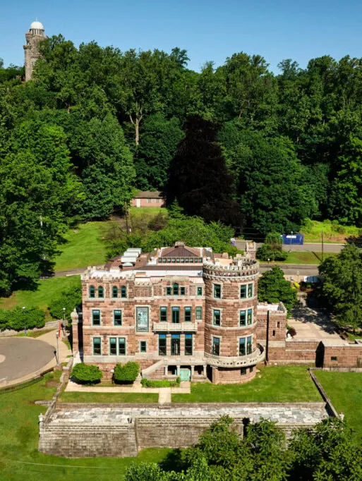 Lambert Castle Museum in Paterson, New Jersey, viewed from above on a sunny day. The historic stone castle sits atop Garret Mountain Reservation, featuring castle-style architecture, landscaped grounds, and wooded hills surrounding the property. A stone observation tower rises in the background above the treeline.