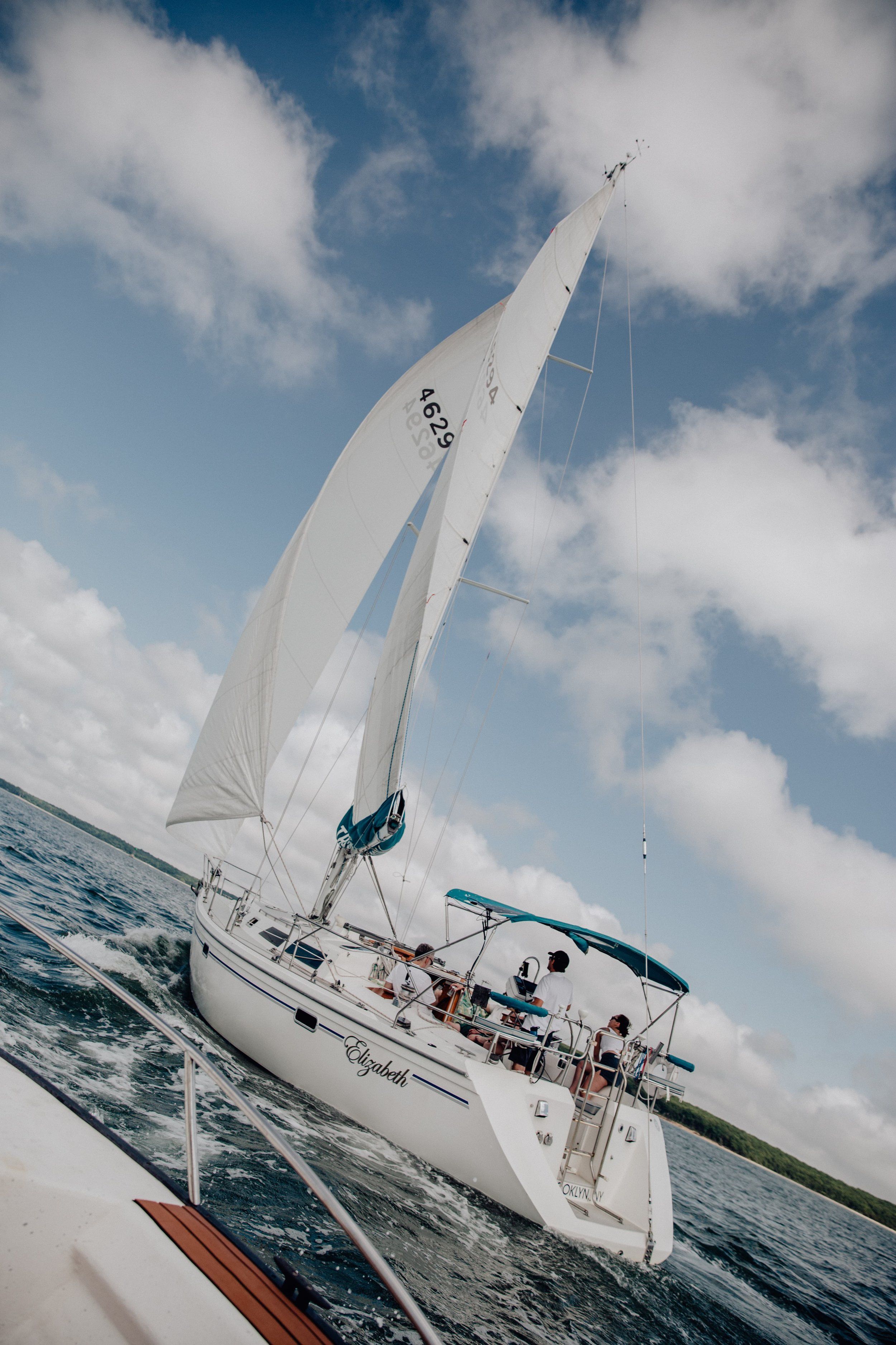 White sailboat cruising across open water near Port Monmouth, New Jersey, with its sails fully raised beneath a bright blue sky filled with large white clouds.