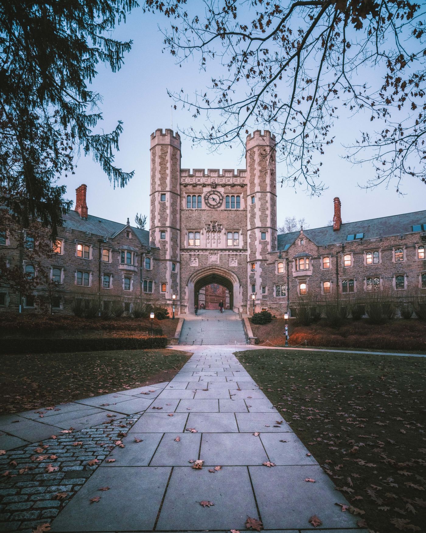 Historic Princeton University campus in Princeton, NJ featuring a Gothic-style stone archway and clock tower, with a cobblestone walkway leading through the courtyard and scattered autumn leaves under an overcast sky.