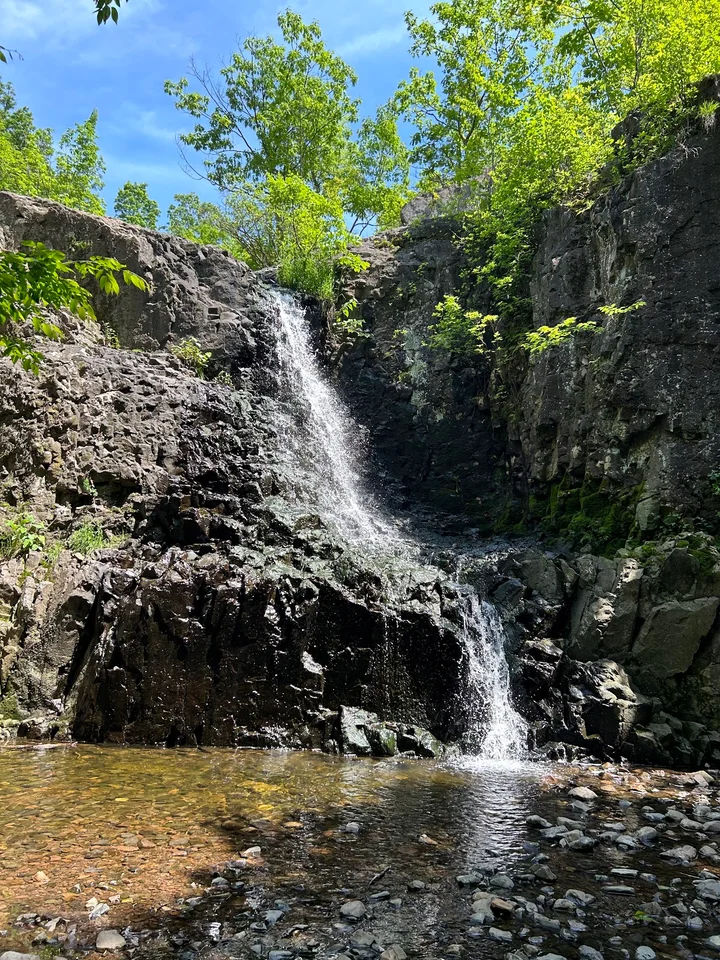 Waterfall at South Mountain Reservation near Roseland, New Jersey, featuring cascading water flowing over rugged rock formations into a shallow pool, surrounded by forest and lush greenery.