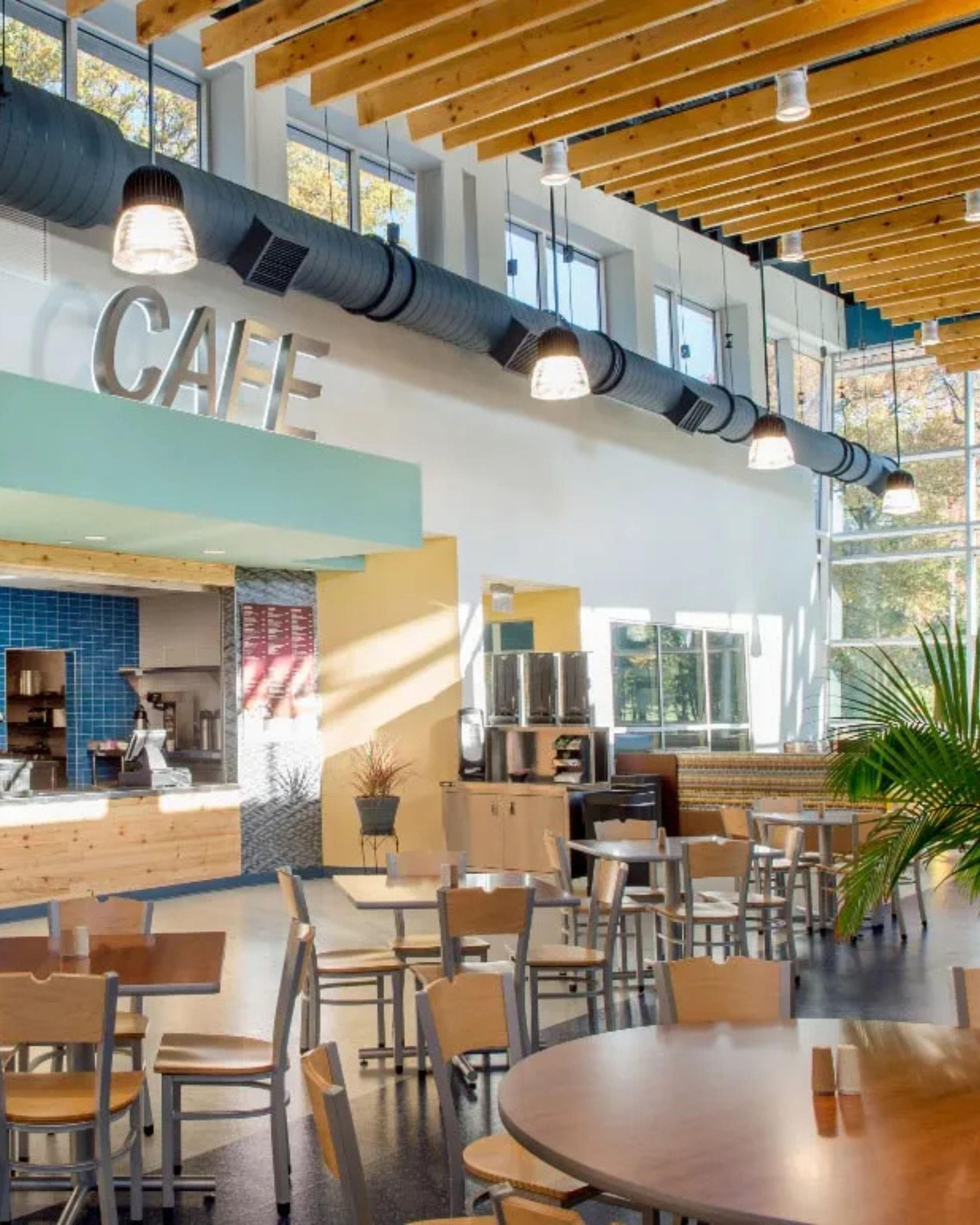 Modern café interior within the Warinanco Park building in Roselle, New Jersey, featuring high ceilings with exposed wood beams, large windows, pendant lighting, and casual seating areas.