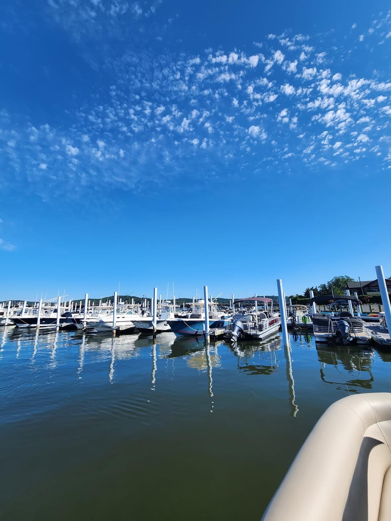 Marina in Rumson, New Jersey, with rows of motorboats and pontoon boats docked along white pilings on calm water beneath a vivid blue, cloud-filled sky.