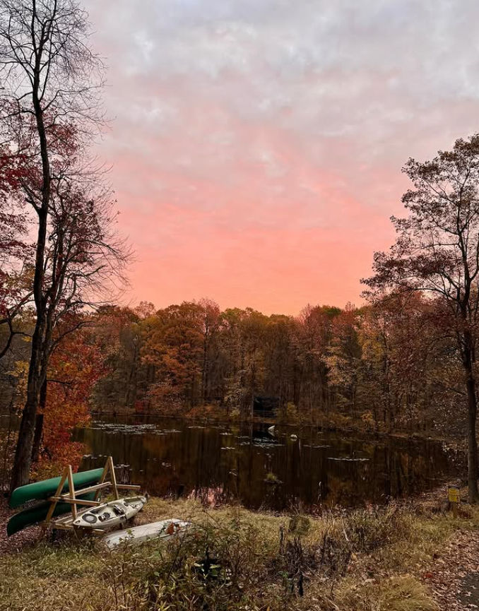 Peaceful pond in Tenafly, NJ surrounded by colorful autumn foliage at sunset, with canoes resting along the shoreline and pink clouds reflecting across the calm water.