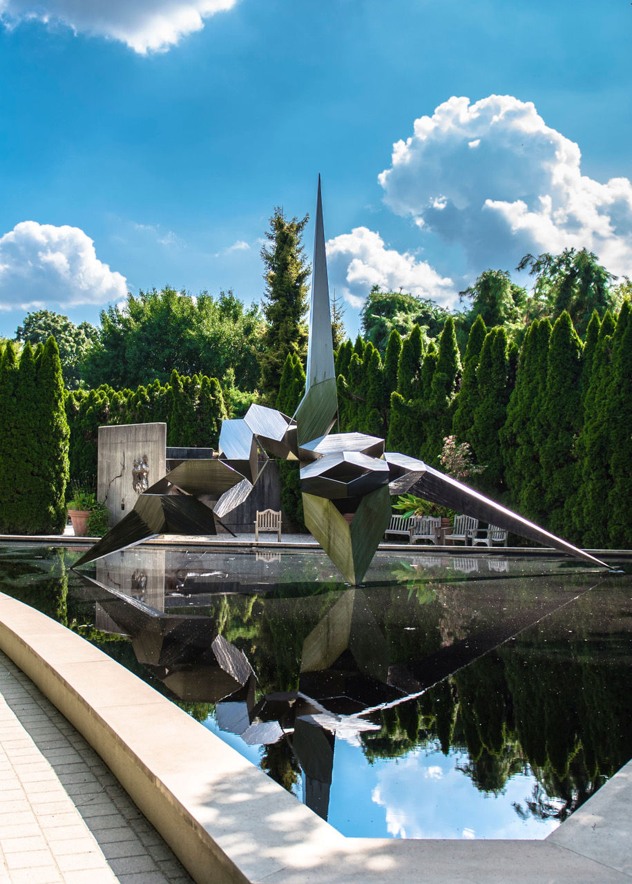 Modern abstract metal sculpture reflected in a still reflecting pool at Grounds For Sculpture in Trenton, New Jersey. The angular stainless-steel artwork rises above the water, surrounded by manicured evergreen hedges, garden pathways, and outdoor seating beneath a bright blue sky with large white clouds. The sculpture’s geometric design creates a striking mirrored reflection across the surface of the pool.