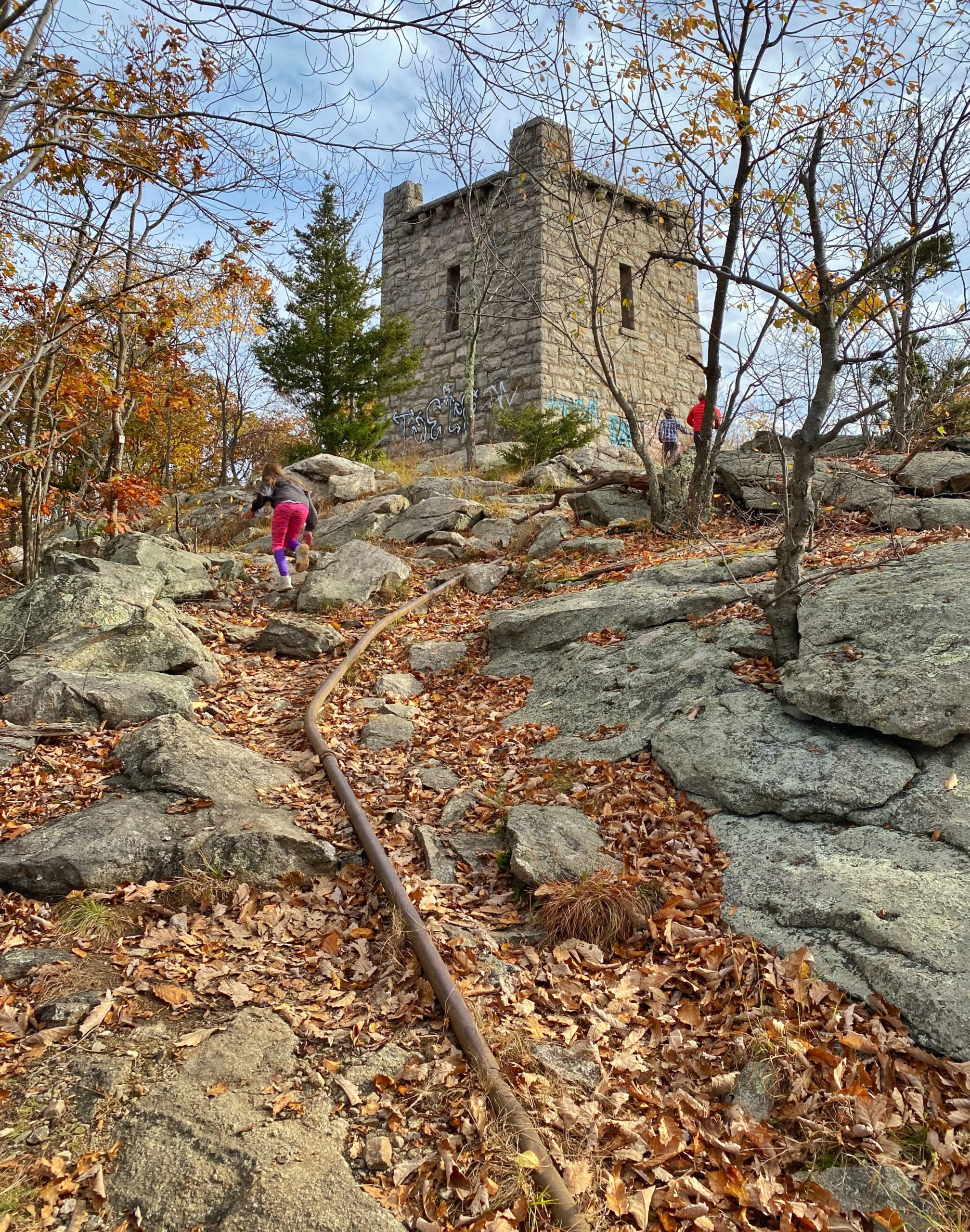 Van Slyke Castle ruins in Ramapo Mountain State Forest near Wanaque, New Jersey, featuring the stone tower surrounded by a rocky hiking trail, bare trees, and fallen autumn leaves.