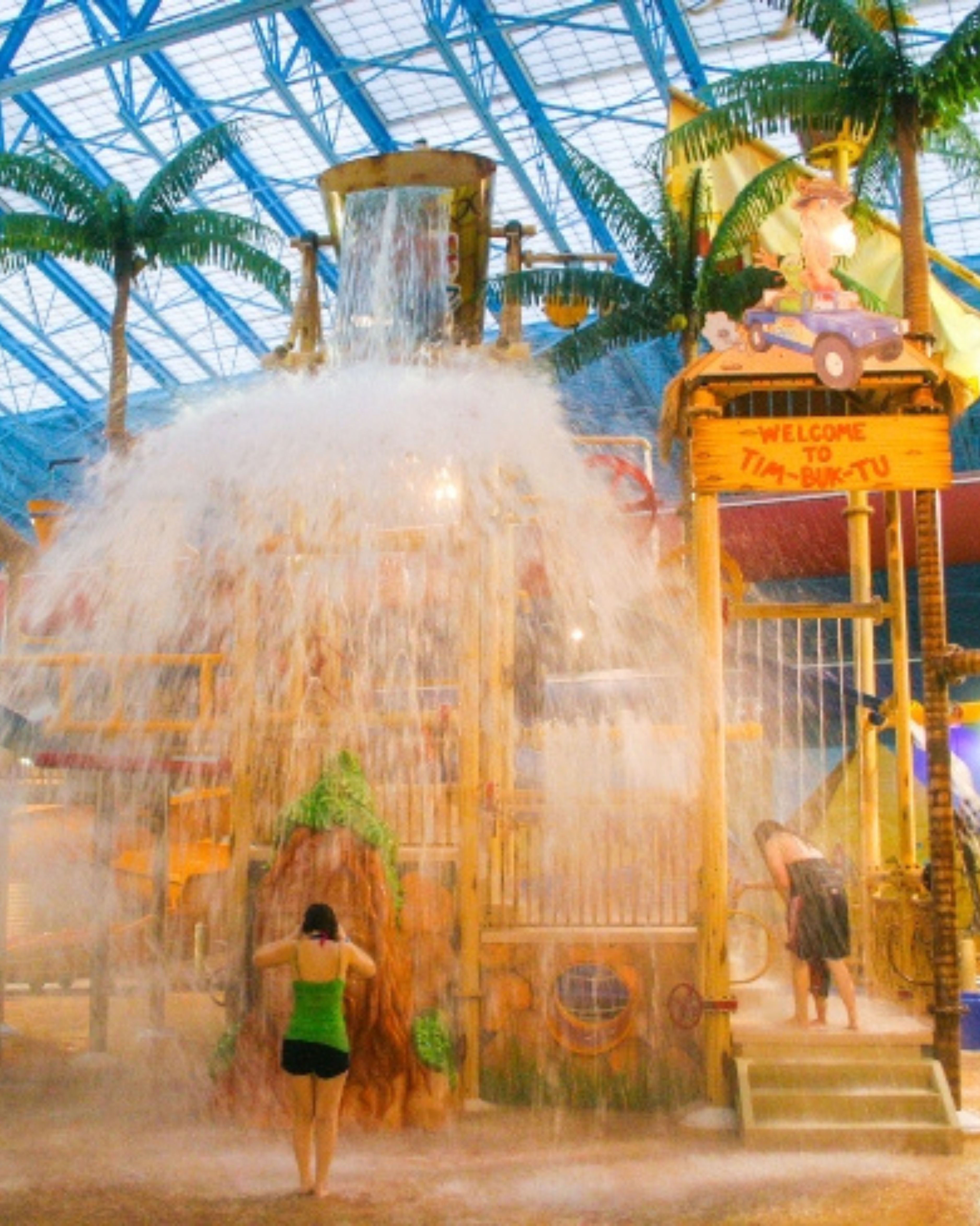 Indoor view of a family water park in West Berlin, New Jersey, featuring a large splash play structure with cascading water, palm tree decor, and visitors enjoying the attraction.