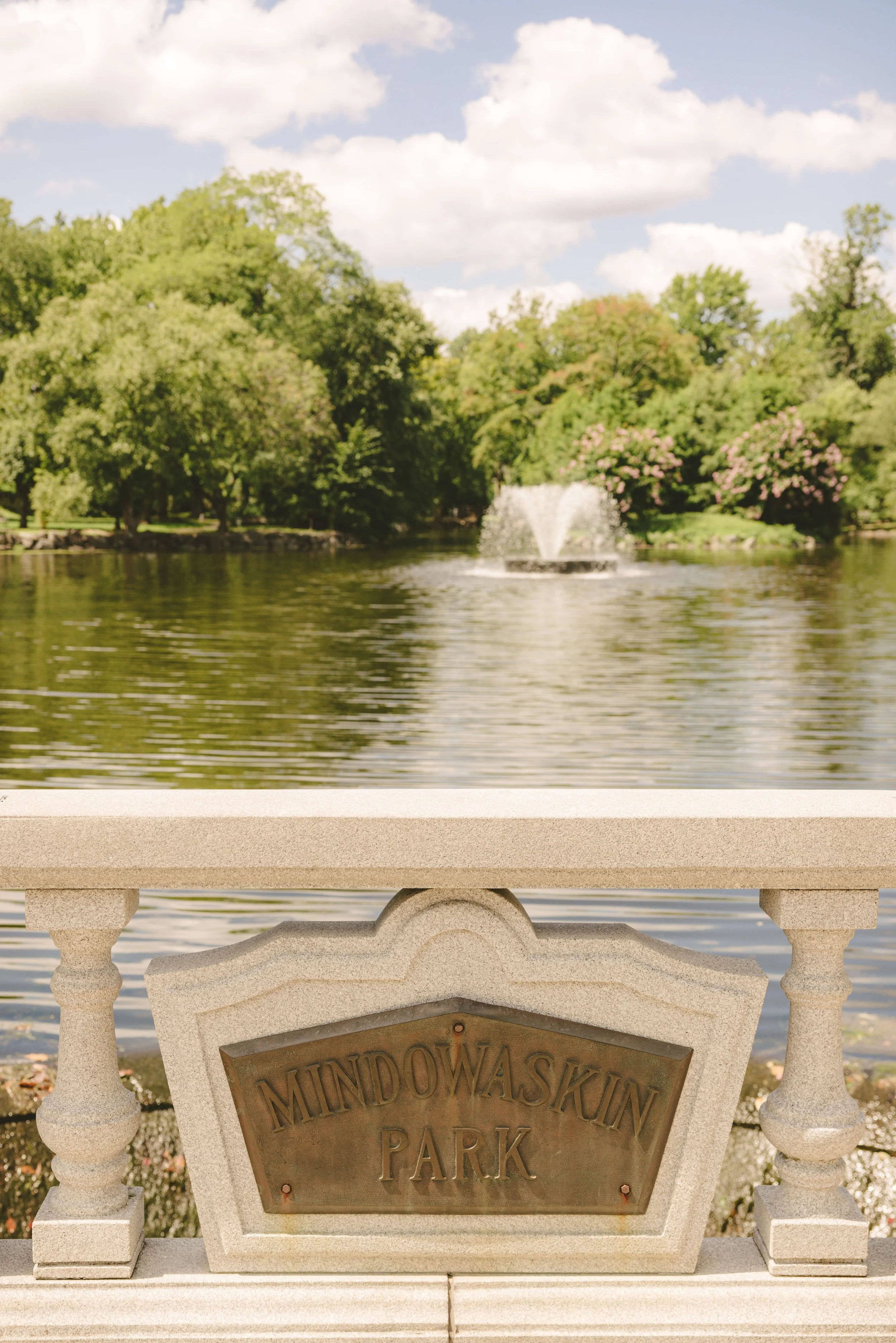 View of Mindowaskin Park in Westfield, New Jersey, featuring a peaceful pond with a fountain, lush greenery, and the park’s stone overlook sign in the foreground.