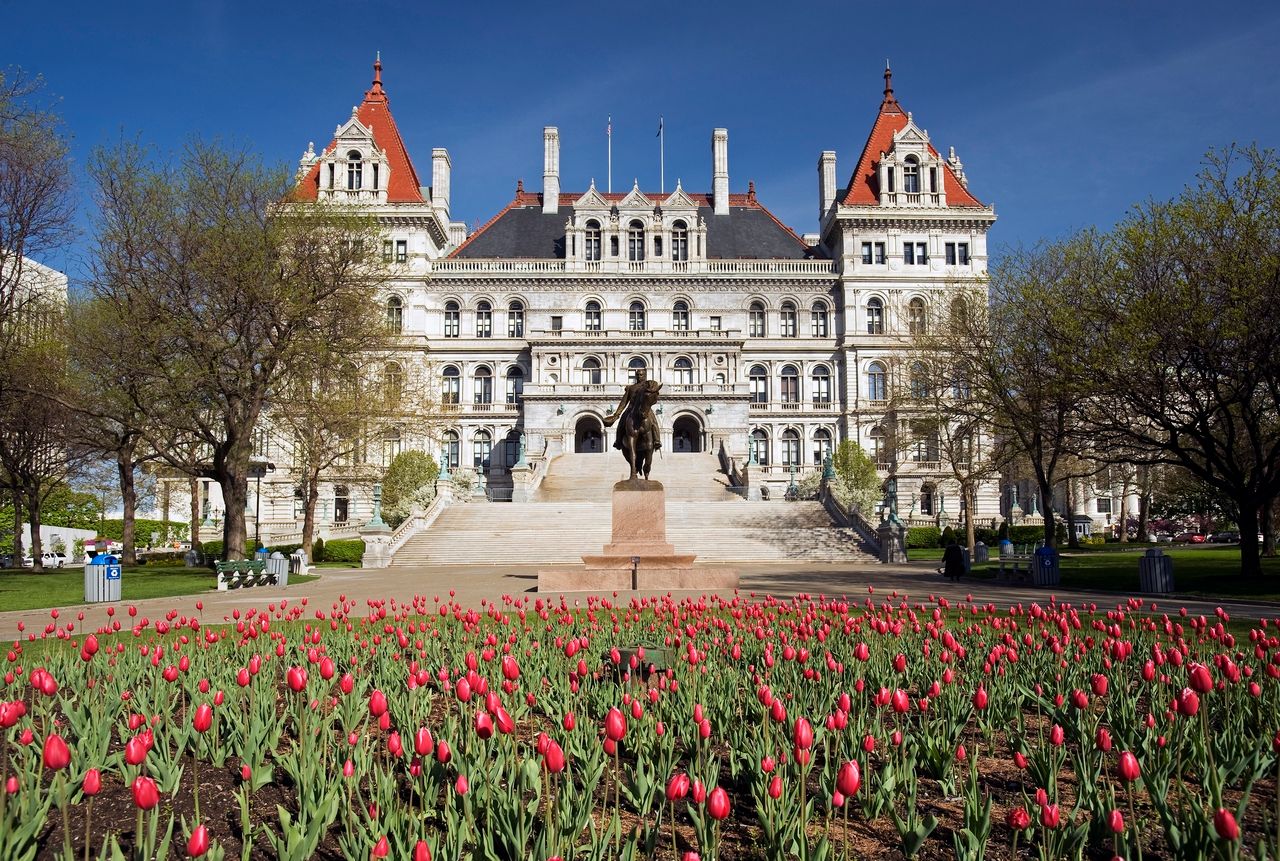 Front view of the New York State Capitol building in Albany, New York, featuring its grand historic architecture with ornate stone detailing and red-roofed towers, a central staircase leading up to the entrance, and a statue in the foreground surrounded by a vibrant bed of blooming red tulips and landscaped grounds.
