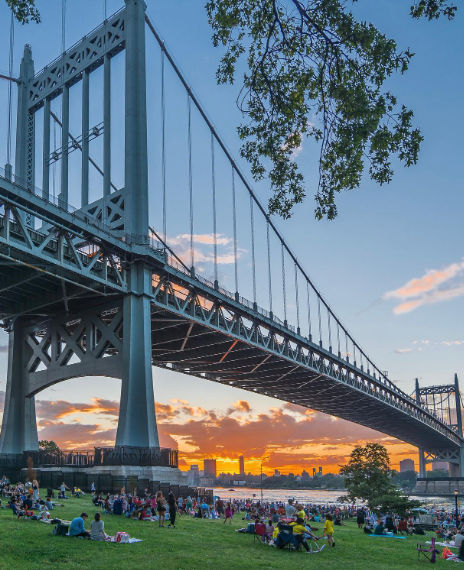 View from Astoria Park in Astoria, New York, showcasing the Robert F. Kennedy (Triborough) Bridge stretching across the East River as people gather on the lawn during sunset.