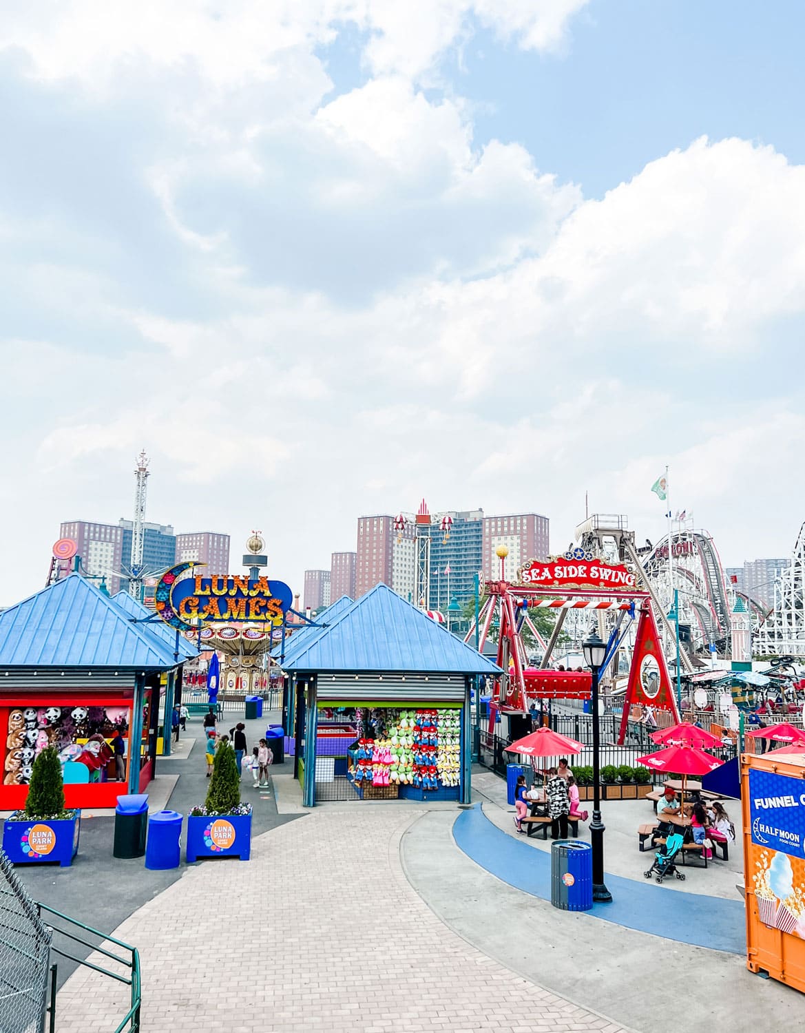 Visitors explore Luna Park in Brooklyn, New York, featuring colorful game booths, amusement rides, and family-friendly attractions along the famous Coney Island boardwalk. Bright signage, carnival games, and classic amusement park rides create a lively seaside atmosphere.