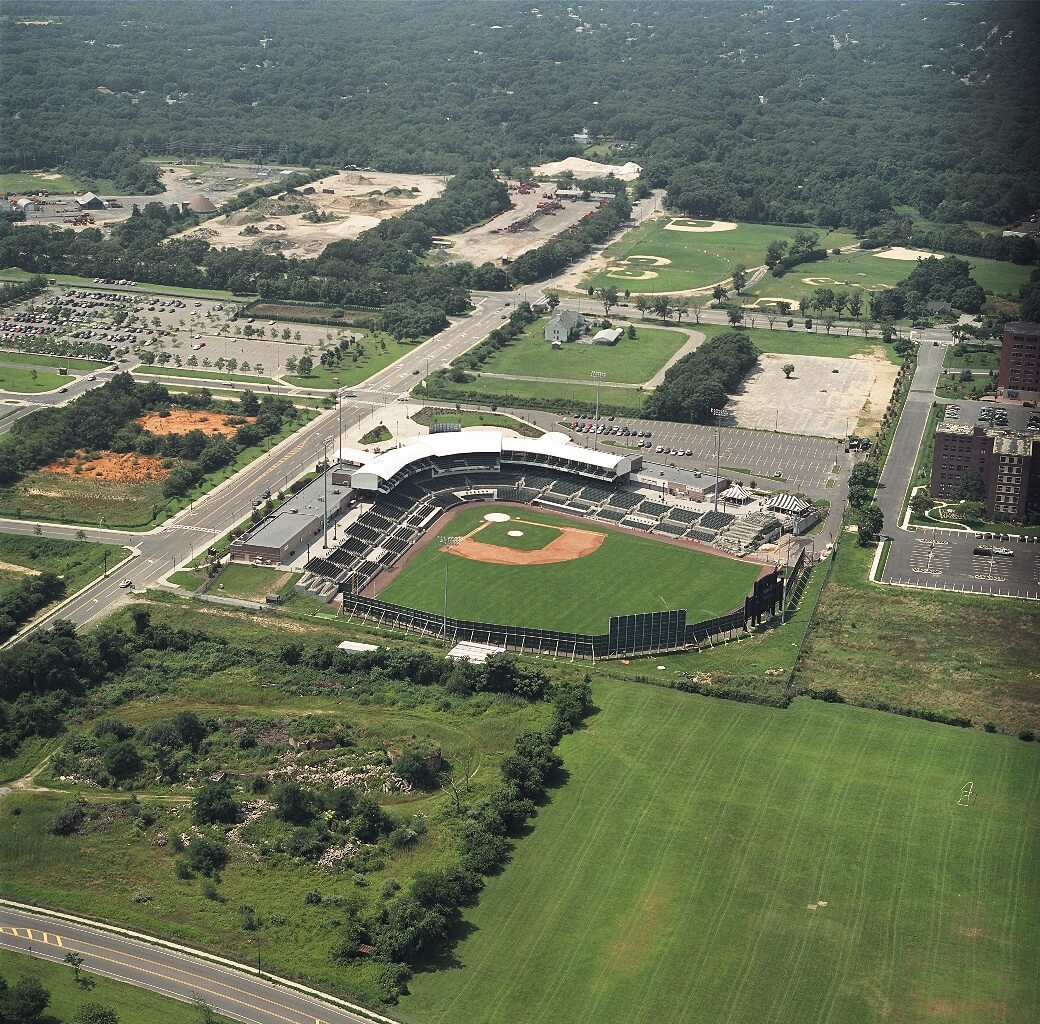 Aerial view of the Long Island Ducks’ home stadium in Central Islip, NY, featuring the baseball field, grandstands, and surrounding parking areas, with nearby roads, open fields, and wooded landscapes stretching into the distance.