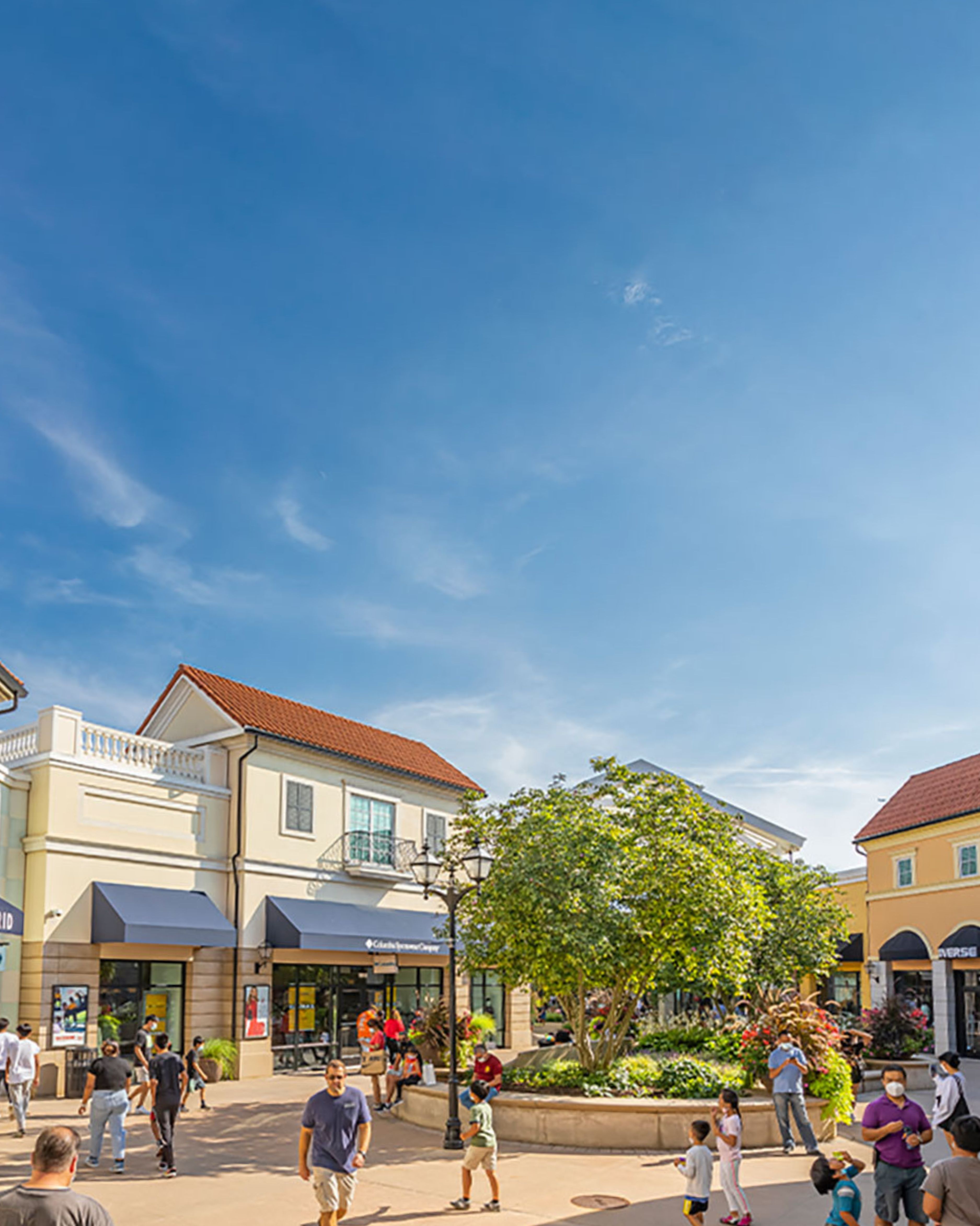 Outdoor view of a busy shopping plaza in Deer Park, New York, featuring storefronts, landscaped walkways, and visitors enjoying a sunny day at Tanger Outlets.