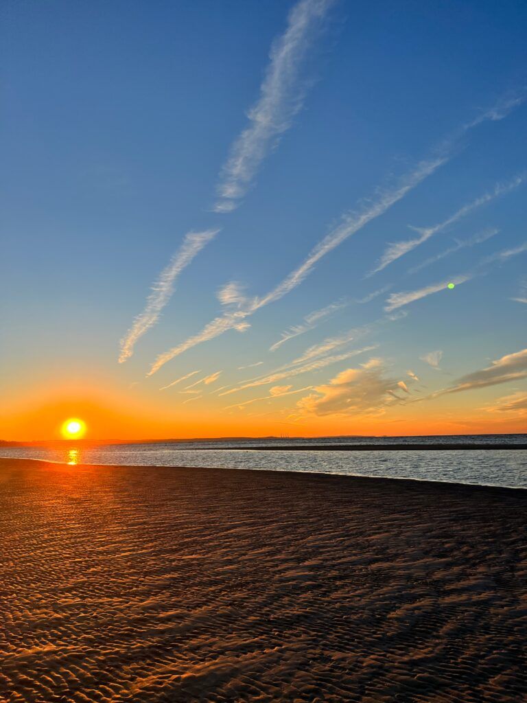 Scenic sunset view at West Meadow Beach near East Setauket, New York, featuring calm shoreline waters, rippled sand, and a colorful sky streaked with clouds at dusk.