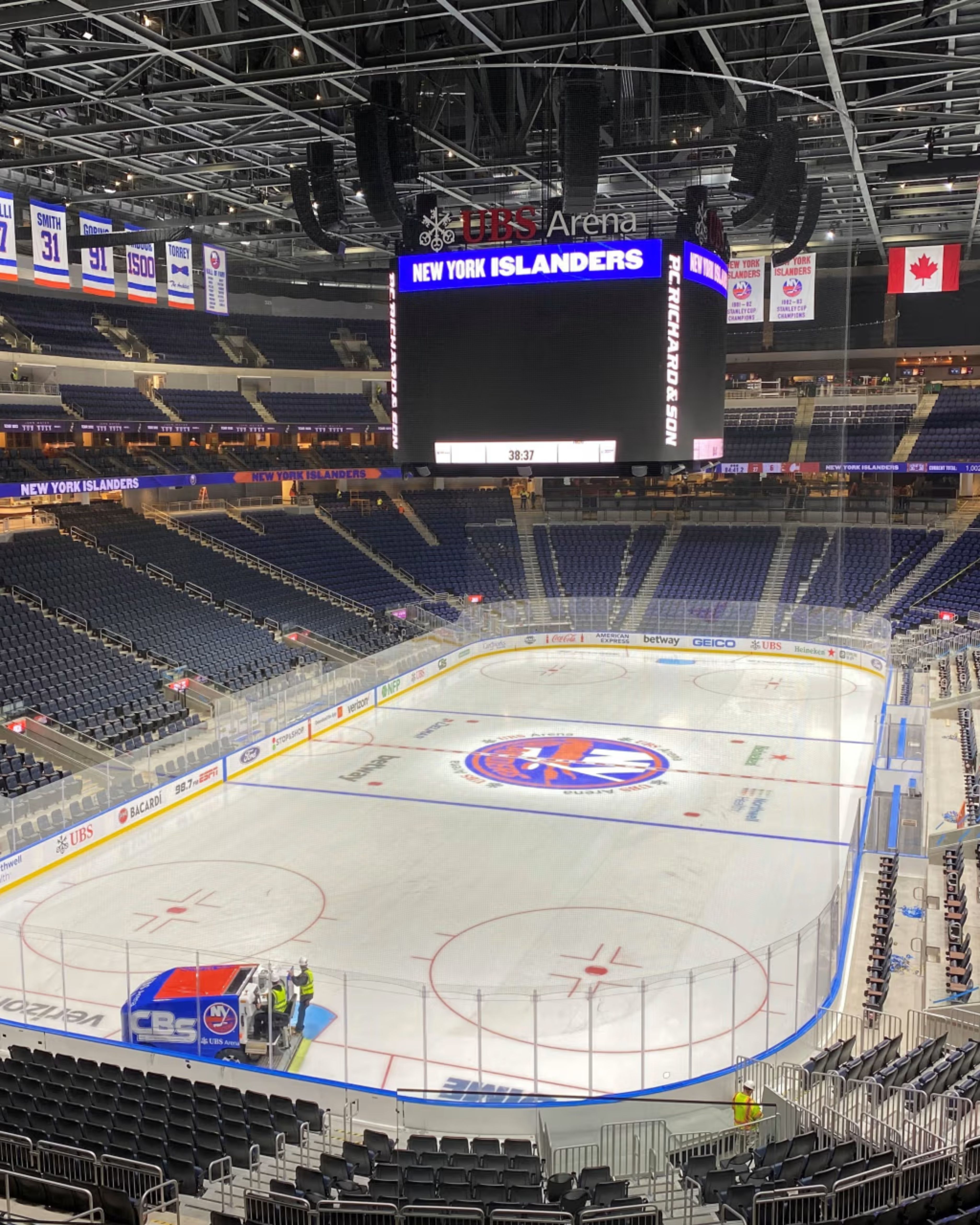 Interior view of UBS Arena in Elmont, New York, featuring the New York Islanders hockey rink, large center-hung scoreboard, surrounding arena seating, championship banners, and bright overhead lighting before an event.
