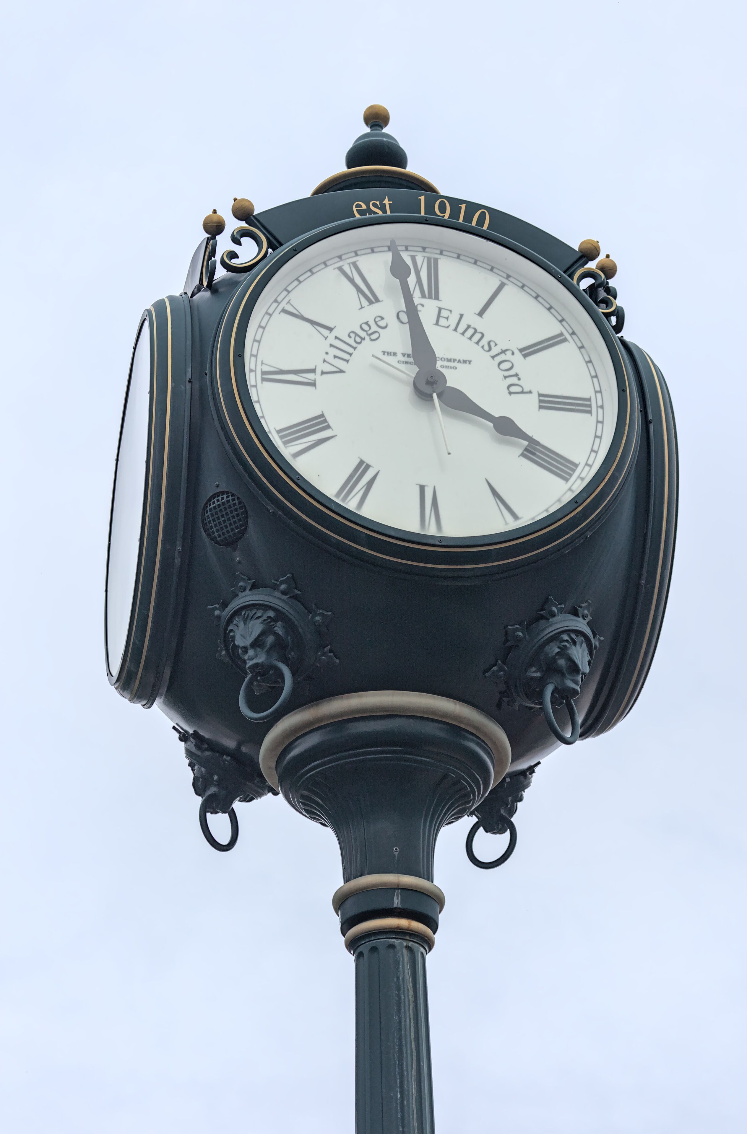Close-up view of the Village of Elmsford historic street clock, established in 1910, featuring Roman numerals, decorative black metal detailing, gold accents, and an ornate pedestal.