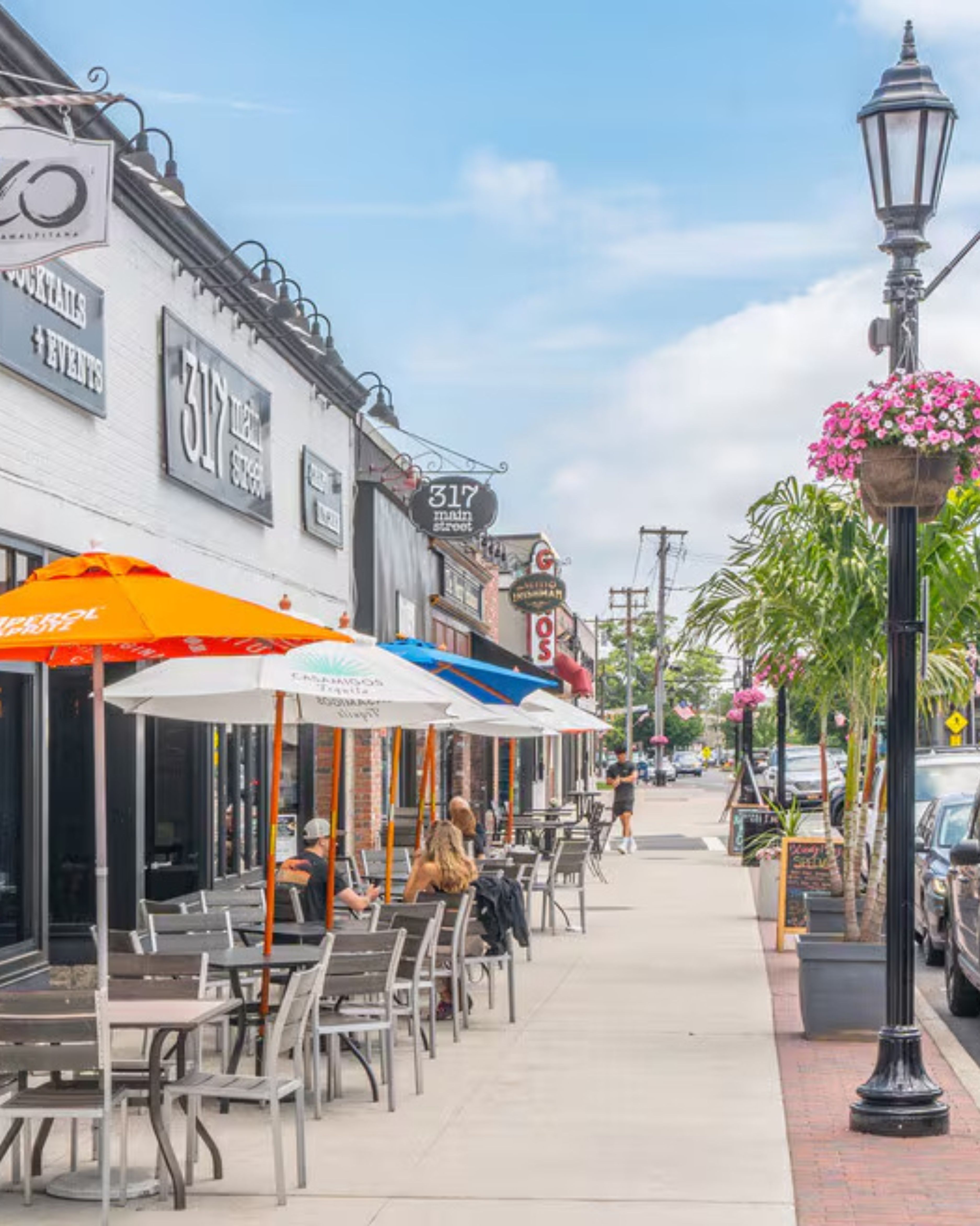 Outdoor dining scene along Main Street in Farmingdale, NY, featuring local restaurants with patio seating, colorful umbrellas, storefront signage, and a walkable sidewalk lined with street lamps and flowers.