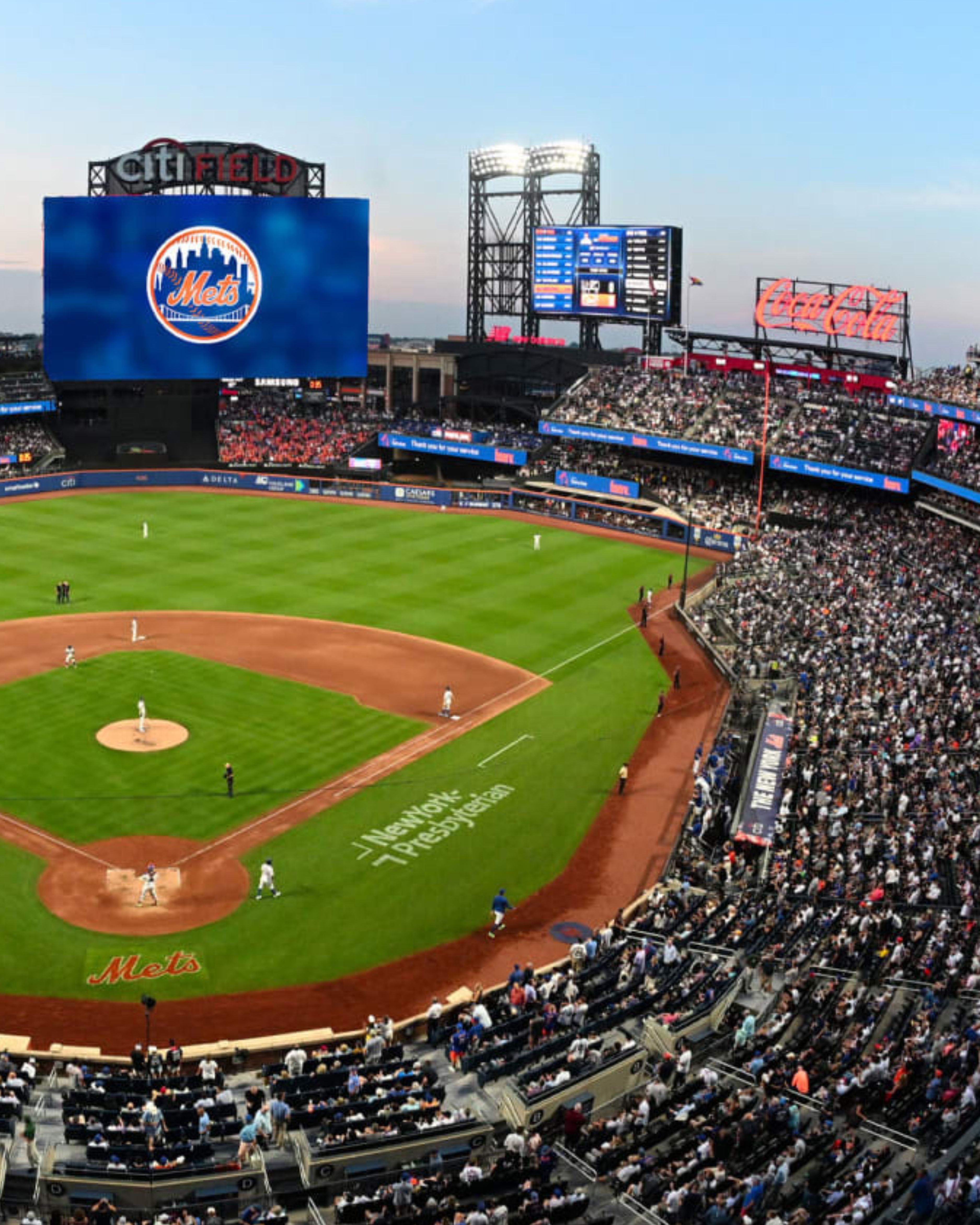 Crowd-filled view of Citi Field in Flushing, NY during a New York Mets game, featuring the baseball field, scoreboard, and packed stadium seating under bright lights