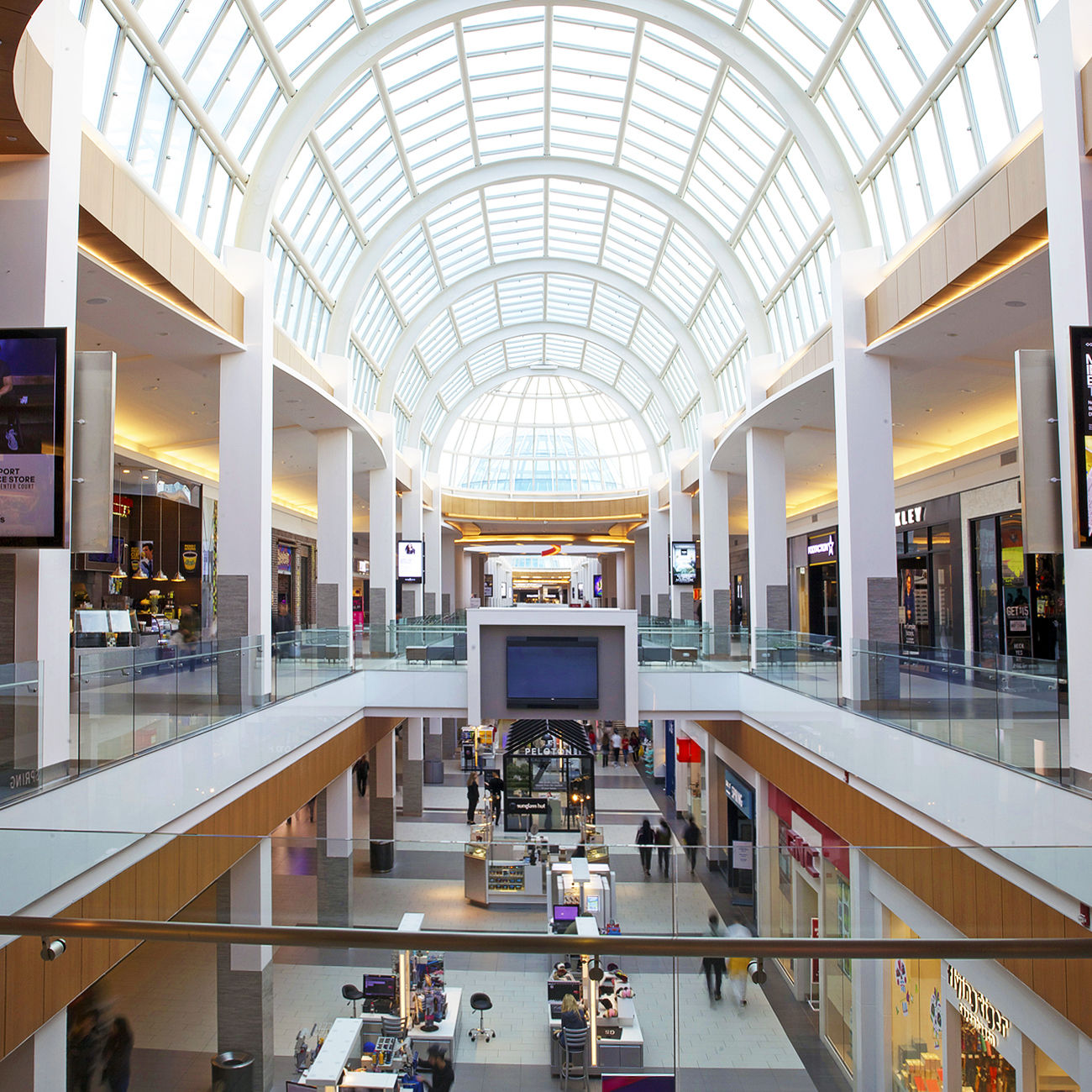 Bright, modern interior of Roosevelt Field Mall in Garden City, NY, featuring a large glass atrium ceiling, multiple shopping levels, and a variety of retail stores and kiosks with shoppers walking throughout.