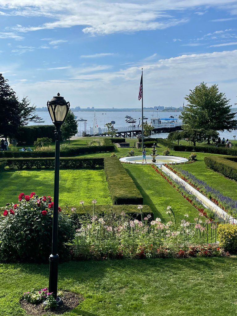 Steppingstone Park in Great Neck, New York featuring manicured gardens, a central fountain, waterfront views of the Long Island Sound, and a pier with boats in the distance.