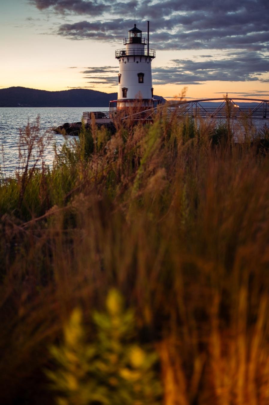 Historic white lighthouse with a black lantern room standing along the Hudson River in Westchester County, New York. A metal pedestrian bridge connects to the lighthouse, while tall golden grasses, distant wooded hills, and a dramatic sunset sky frame the waterfront scene.