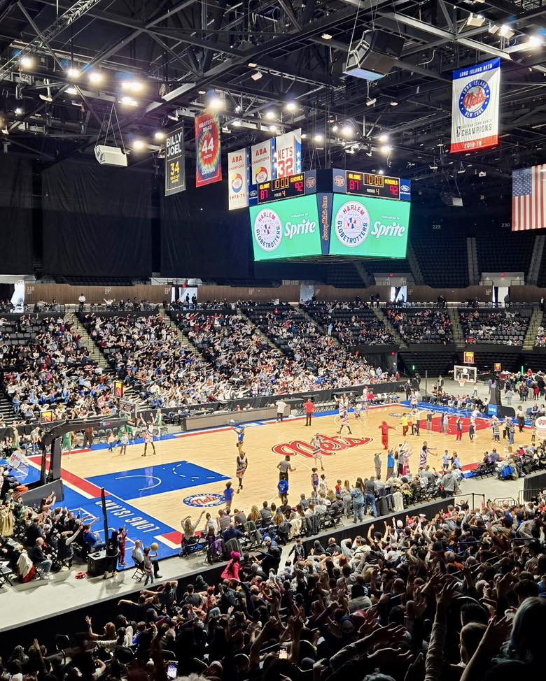 Interior view of Nassau Coliseum in Hempstead, NY during a live basketball event, featuring a packed crowd, illuminated scoreboard, and players on the court creating an energetic game-day atmosphere