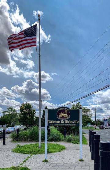 Welcome to Hicksville sign beneath an American flag in Hicksville, New York, featuring landscaped greenery, a pedestrian walkway, nearby streets, and a bright blue sky with scattered clouds.