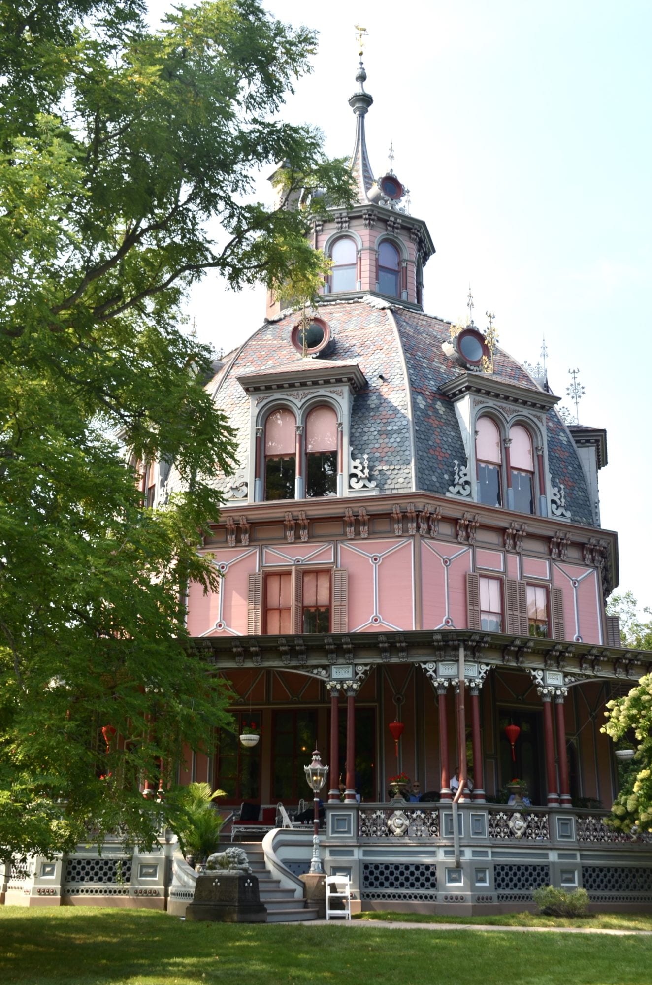 Historic Lyndhurst Mansion near Irvington, New York, featuring an ornate Victorian-style exterior with pink walls, decorative patterned roofing, arched windows, a tall central spire, and an elaborate covered porch surrounded by mature green trees and landscaped grounds.