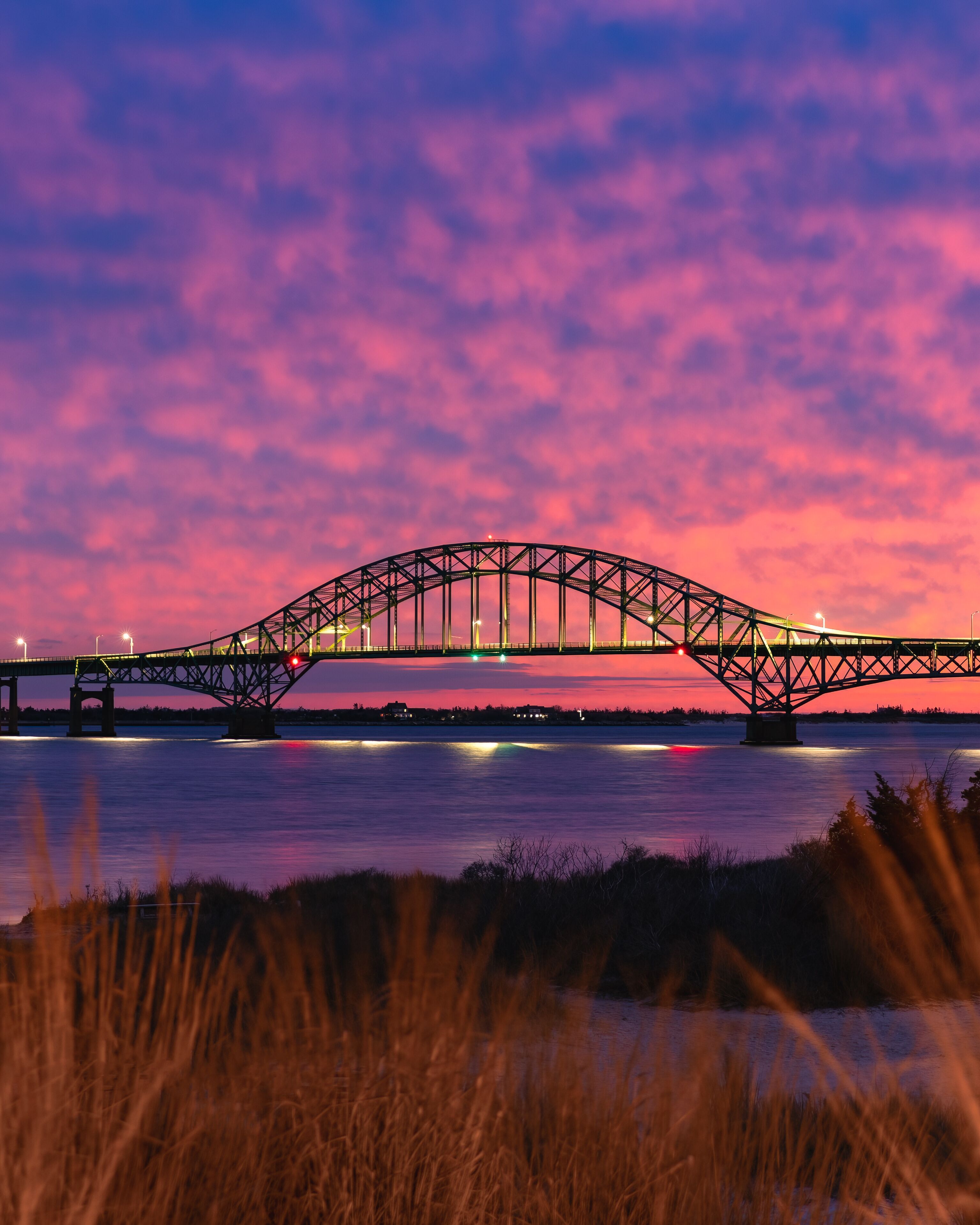 Sunset view of the Robert Moses Causeway Bridge near Islip, New York, featuring the illuminated steel arch bridge stretching across calm coastal water beneath a vibrant pink and purple evening sky.