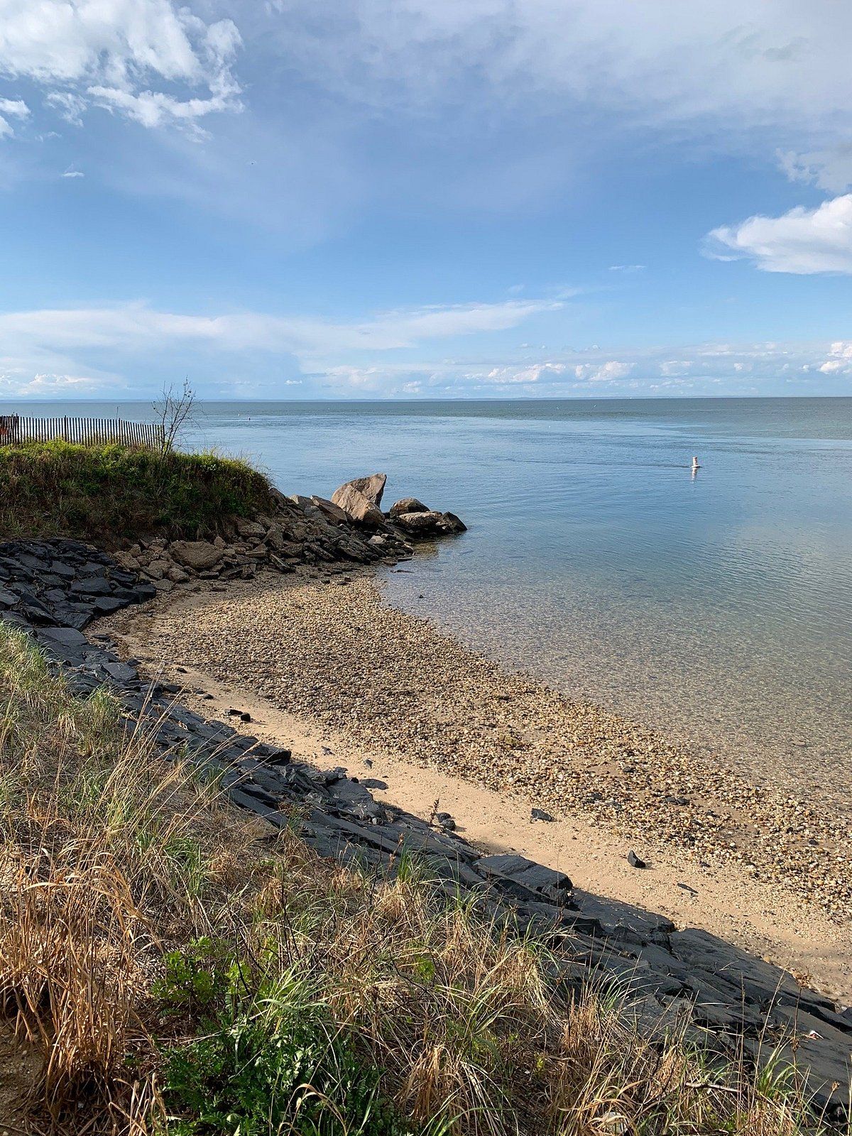 Rocky shoreline along the Long Island Sound in Kings Park, NY with calm blue water, scattered stones, and a peaceful coastal view under a lightly clouded sky