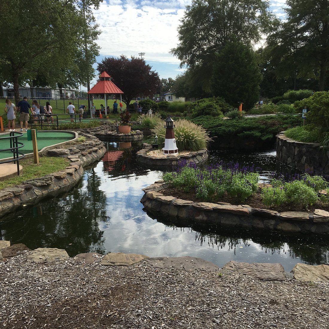 A landscaped mini golf course in Levittown, New York, featuring a small pond with stone edging, decorative lighthouse structures, and families enjoying the course surrounded by greenery.