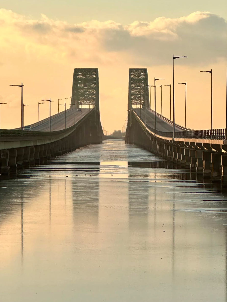 Scenic view of the Robert Moses Causeway bridge near Lindenhurst, NY at sunset, featuring symmetrical spans over calm water with soft golden reflections and Long Island’s coastal landscape in the distance.