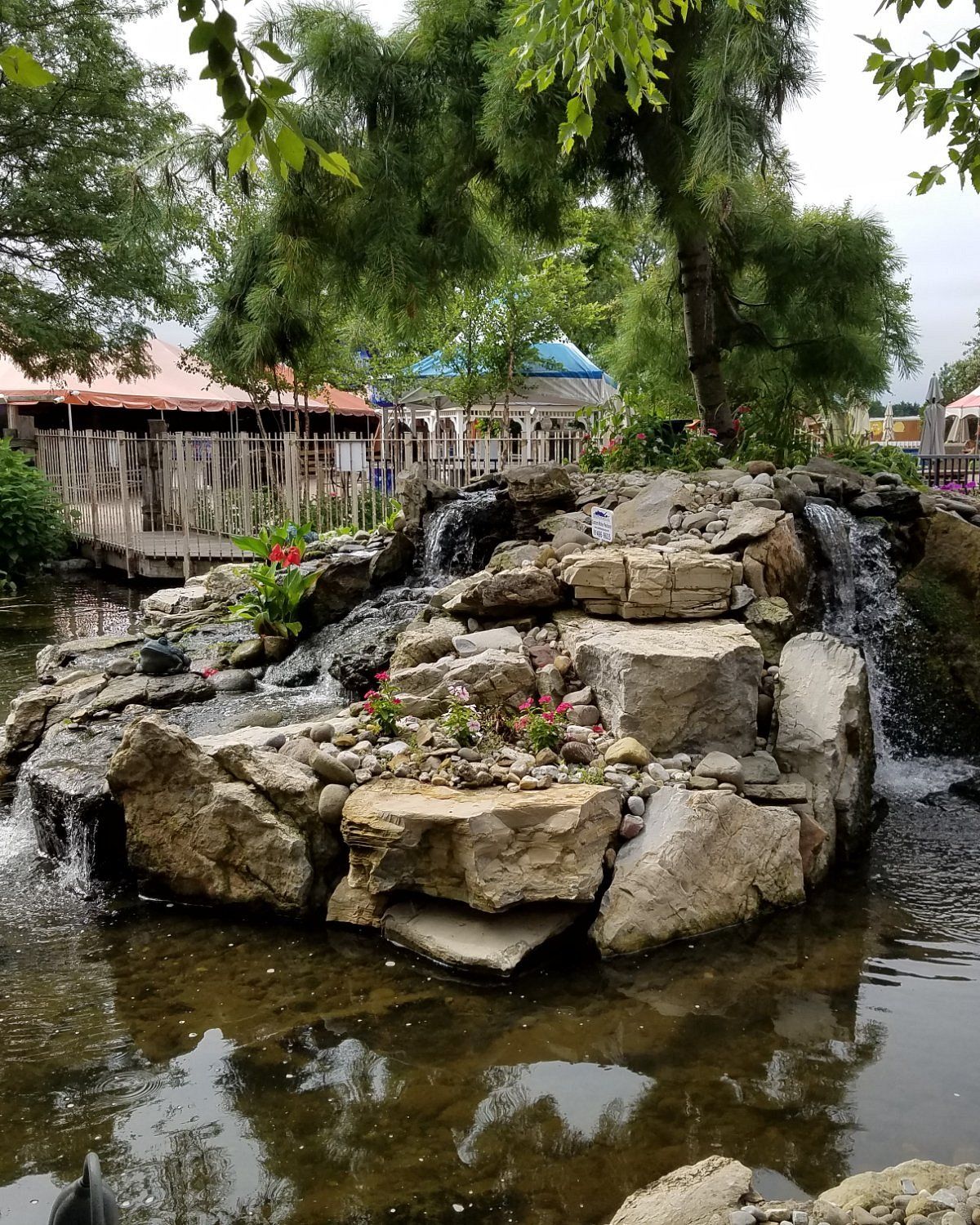 Landscaped outdoor waterfall and rock garden at a recreational property in Melville, New York, featuring cascading water flowing over large natural stones into a pond. Tall pine trees, flowering plants, a wooden pedestrian bridge, and covered outdoor seating areas surround the peaceful garden.