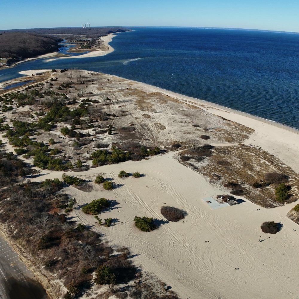Aerial view near Nesconset, New York, featuring the sandy shoreline and coastal waters of Smith Point County Park, with expansive beachscape and surrounding natural landscape under a clear blue sky.
