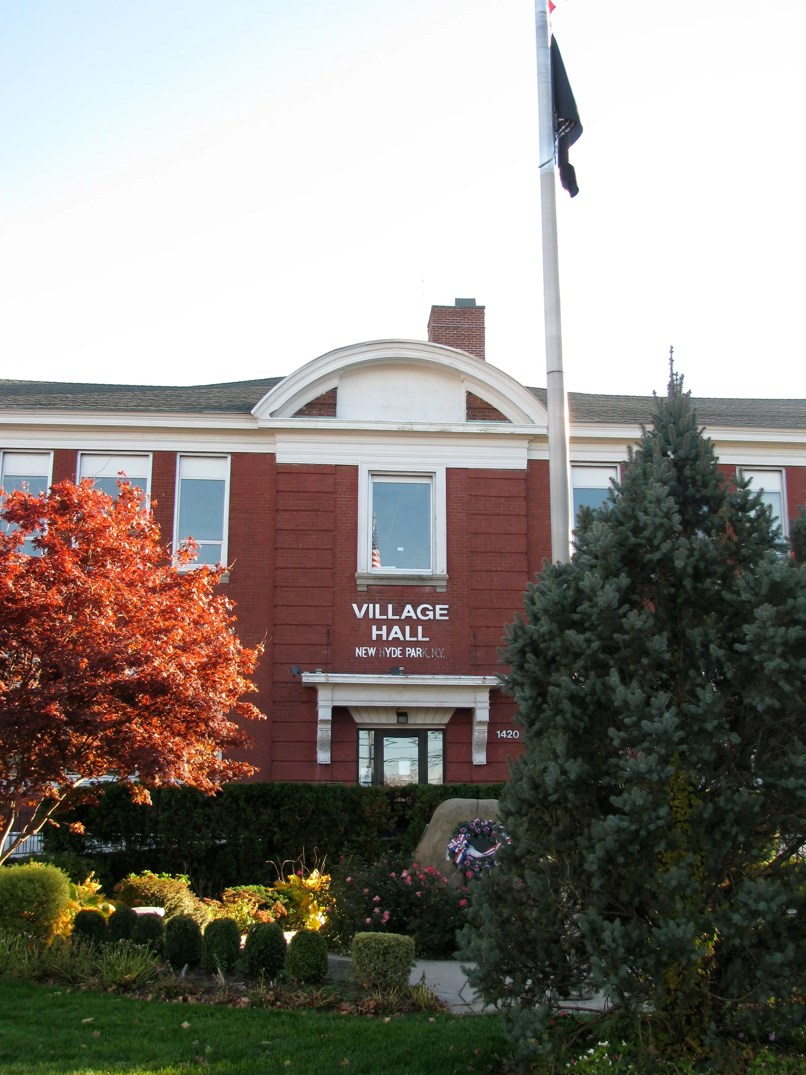 Exterior view of Village Hall in New Hyde Park, New York, featuring a historic red-brick facade with white architectural trim, landscaped gardens, mature trees, a flagpole, and a memorial wreath near the entrance.