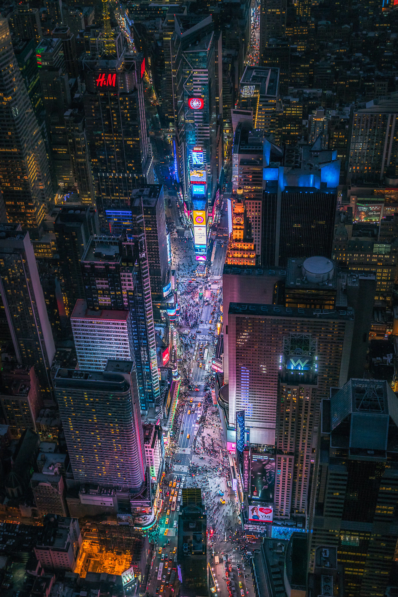 Aerial view of Times Square in New York City at night, showcasing brightly lit billboards, towering skyscrapers, busy streets filled with traffic, and crowds moving through the heart of Manhattan's famous entertainment district.
