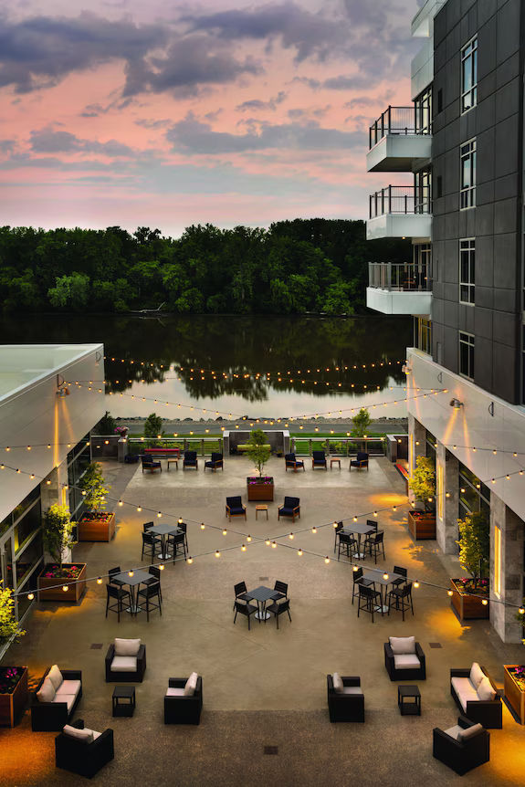 An outdoor waterfront courtyard at Mohawk Harbor in Schenectady, New York, featuring modern apartment buildings, string lights, lounge seating, and dining tables overlooking the Mohawk River. The contemporary gathering space is illuminated at dusk, with reflections on the water and a colorful sunset sky creating a scenic riverside atmosphere.