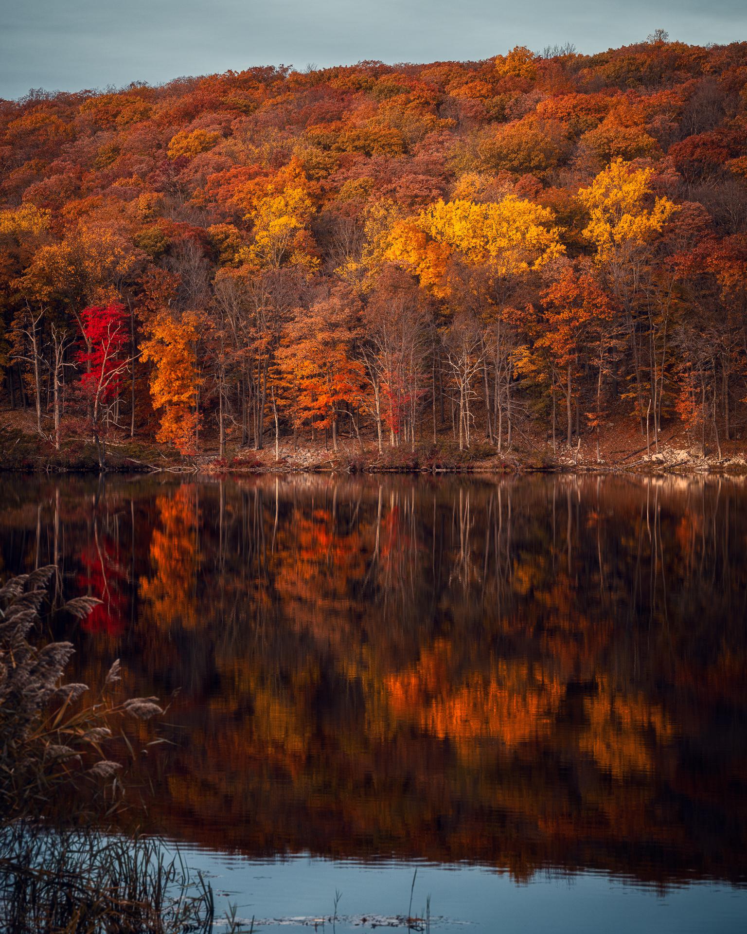 Fall foliage reflected across a peaceful lake near Sloatsburg, New York, with vibrant orange, yellow, and red trees covering the surrounding hillside.