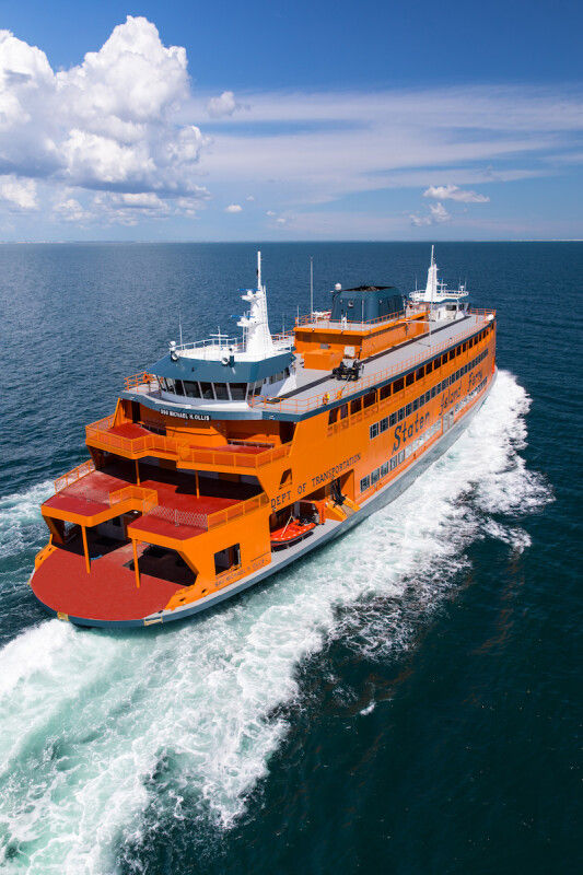 The Staten Island Ferry travels across New York Harbor, leaving a wake behind as it moves through open water under a blue sky. The iconic orange vessel serves as a key transportation link between Staten Island and Manhattan while offering scenic views of the harbor.