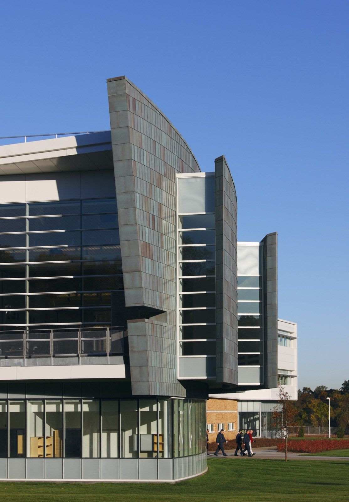 Modern academic building at Stony Brook University in Stony Brook, New York, featuring curved metal exterior panels, expansive glass windows, and contemporary architectural design. Students walk along a landscaped campus pathway beneath a clear blue sky.