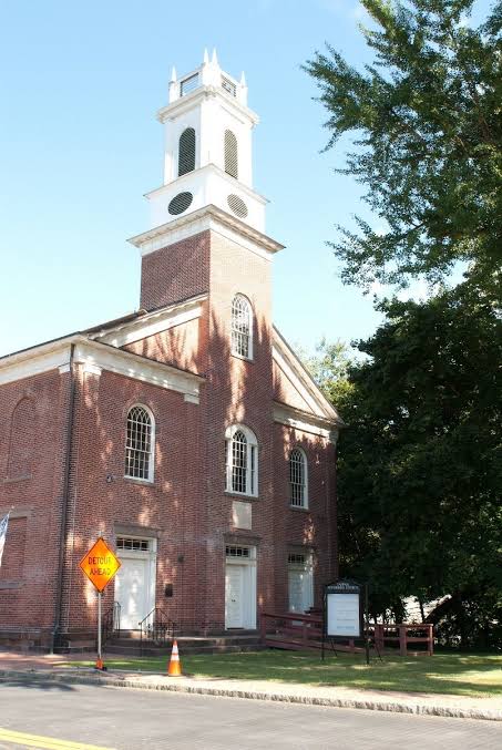 Exterior view of a historic red-brick church in Tappan, New York, featuring tall arched windows, white entrance doors, a prominent white bell tower, and mature trees surrounding the property.