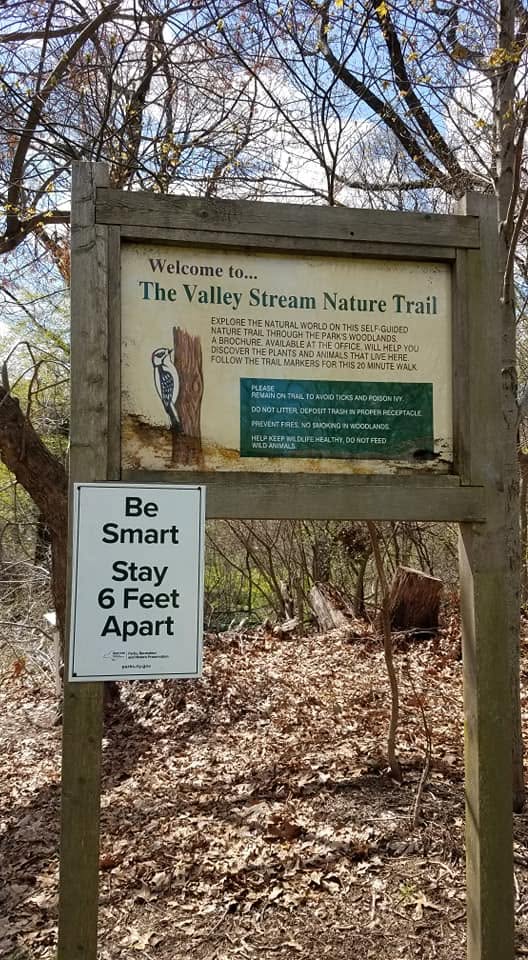 Entrance sign for the Valley Stream Nature Trail in Valley Stream, New York, surrounded by a wooded landscape with mature trees, fallen leaves, and natural vegetation along the trail.