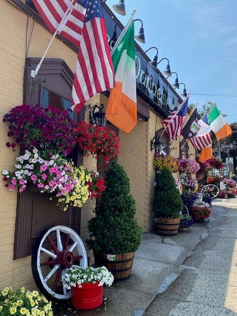 Street view of Dunne’s Pub in White Plains, NY, featuring a charming storefront lined with colorful hanging flowers and multiple flags, creating a lively and welcoming neighborhood atmosphere