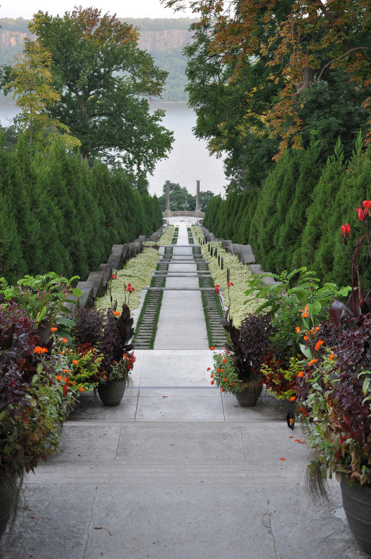 View of Untermyer Gardens in Yonkers, New York, featuring a long, symmetrical stone walkway lined with manicured hedges, vibrant seasonal flowers in planters, and terraced landscaping leading down toward scenic views of the Hudson River in the background.