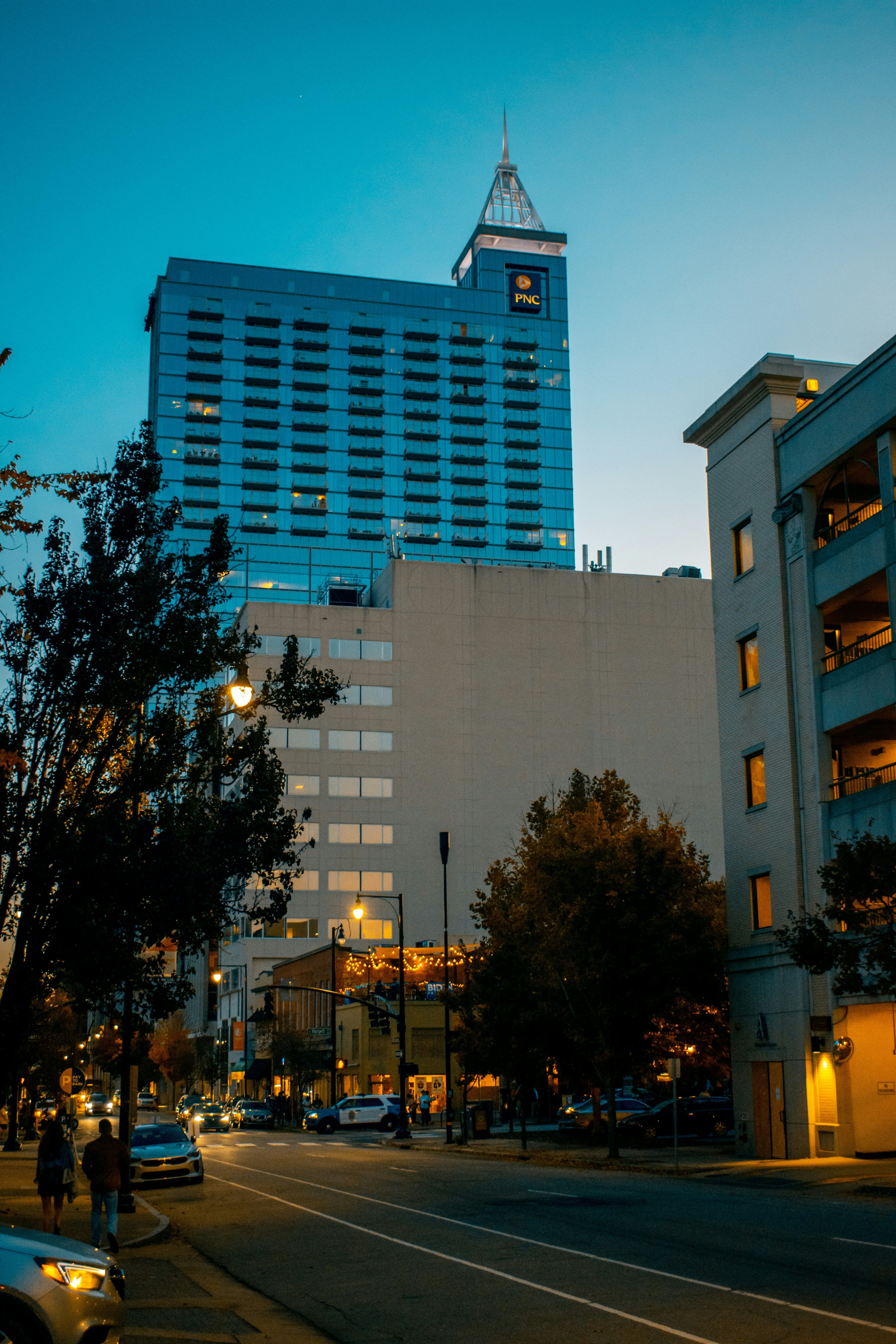 Street-level view of downtown Raleigh, NC with modern glass office buildings, a historic theater marquee, and lush fall trees lining the sidewalk