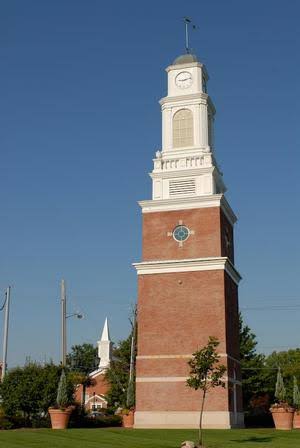 Historic brick clock tower in Strongsville, Ohio, featuring a tall rectangular red-brick base topped with a white decorative clock structure and cupola. The landmark stands against a clear blue sky, with trees, a smaller church steeple, and landscaped grounds visible nearby.