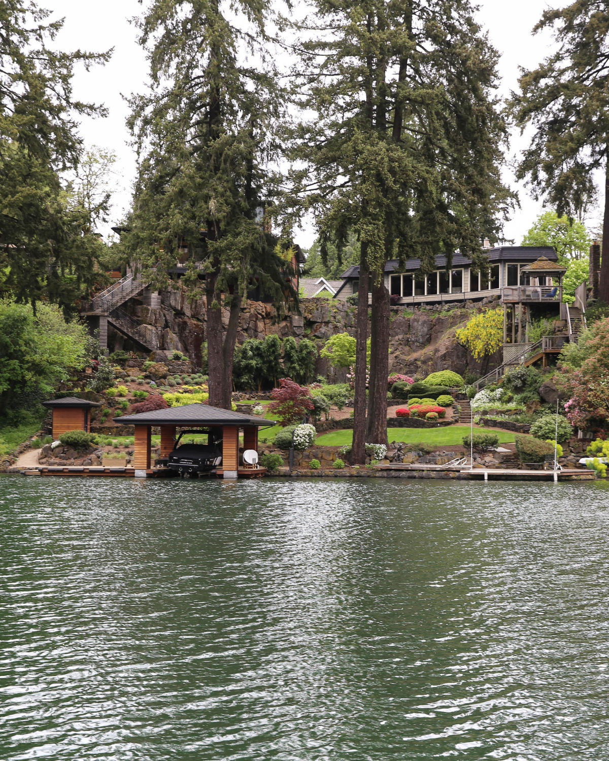 Lakeside view of a terraced hillside garden with colorful flowering shrubs, tall evergreen trees, and a modern home at the top. A small covered boathouse sits at the waterÕs edge in the foreground.